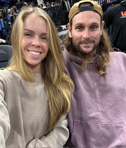 Creative Producer Brittany in a taupe crewneck and Founder Noah in a purple hoodie smiling courtside at a sporting event.