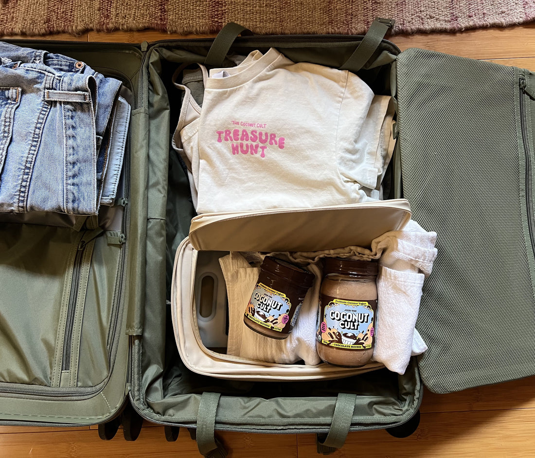 Olive green luggage open on hardwood floor with jeans, t-shirts, and a beige bag holding two jars of Chocolate Mousse Coconut Cult.