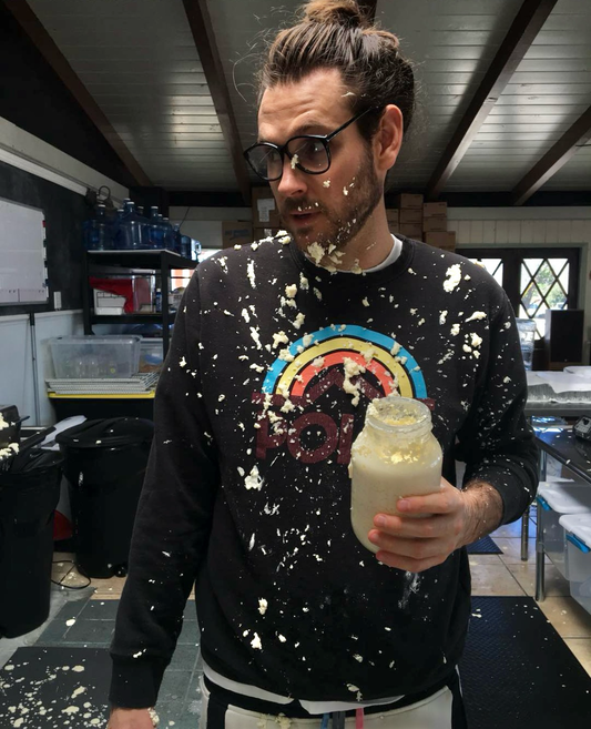 Founder Noah in the kitchen holding an exploded jar of Coconut Cult yogurt, with yogurt splattered on his face, glasses, and shirt.