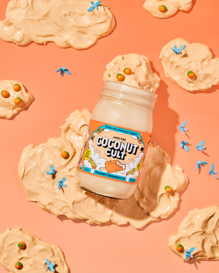 A 16oz jar of Orange Dreamsicle yogurt against a bright orange backdrop with yogurt clouds, mini oranges, and bluebirds