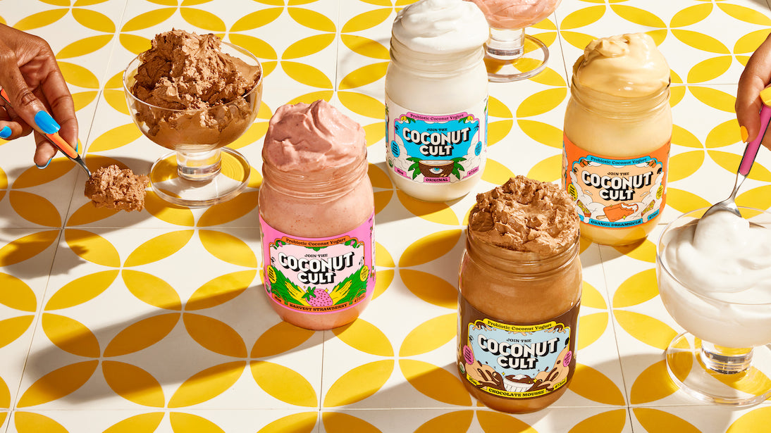 Four 16oz Coconut Cult jars Strawberry, Original, Chocolate, Orange on a lit table between two yogurt-filled bowls