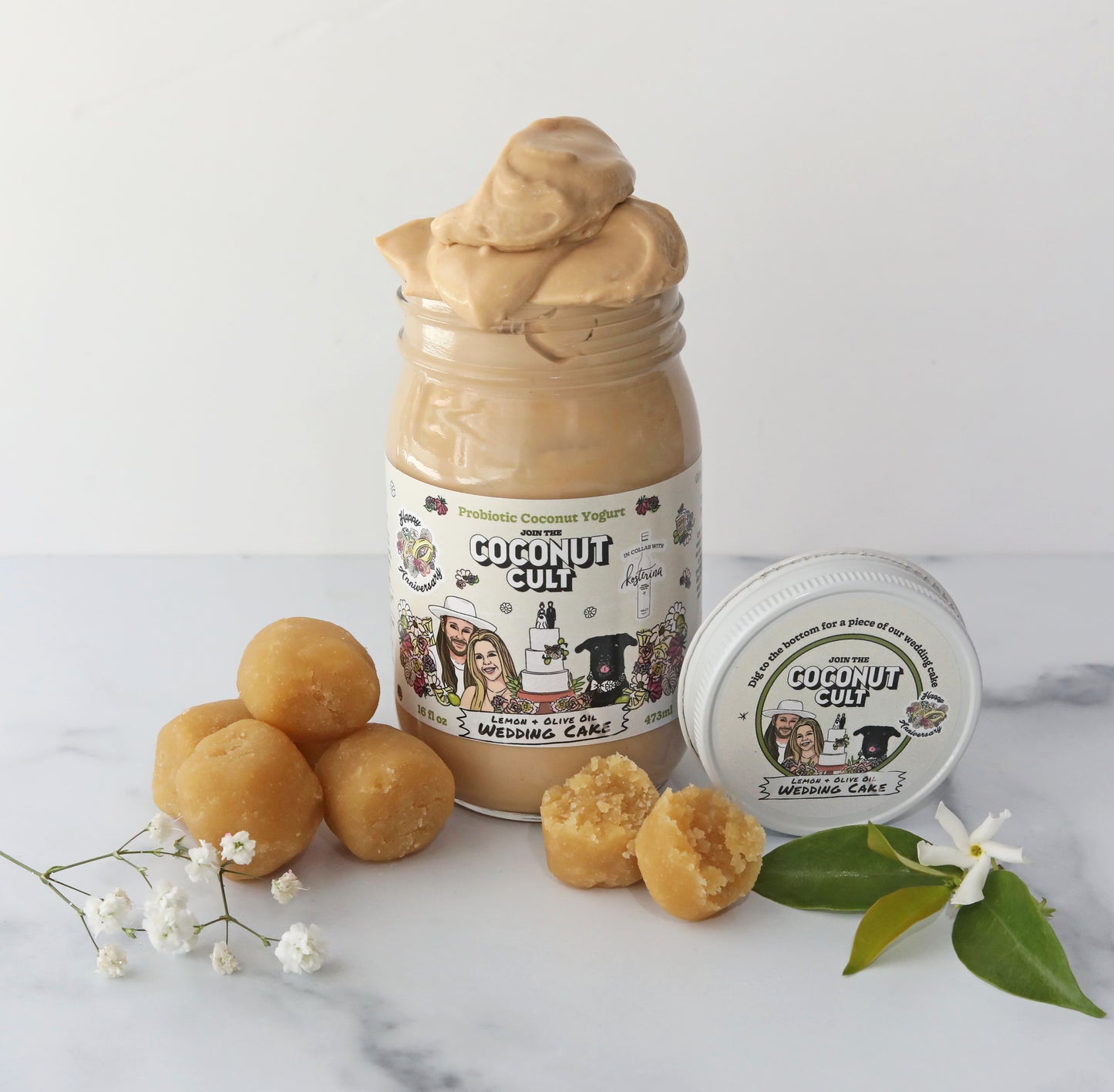 16 oz jar of Lemon & Olive Oil Wedding Cake yogurt with the lid off and yogurt coming out of the top of the jar. The lid is propped up against the jar and there are a couple of leaves and flowers surrounding the jar along with 4 whole cake bites and 1 cake bite that is split in half.